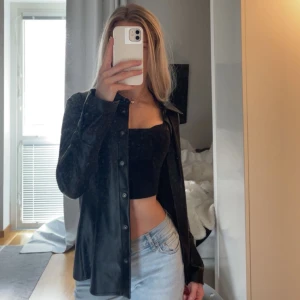 Svart skinnskjorta med knappar - Säljer en svart skjorta i skinnimitation med klassisk krage och knäppning framtill. Skjortan har långa ärmar och en figurnära passform. Perfekt att ha för sig själv eller över en snygg topp! 