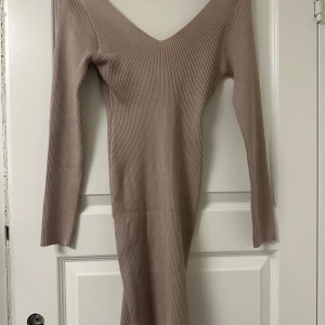 Beige ribbad långklänning med v-ringning - Säljer en beige ribbad långklänning med djup v-ringning och långa ärmar. Klänningen har en figurnära passform och är tillverkad i ett stretchigt material som sitter snyggt på kroppen. Perfekt för dig som gillar stilrena och enkla plagg.