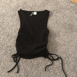 Svart ribbad linnetopp från Divided H&M - Säljer en svart ribbad linnetopp från Divided H&M. Toppen har knytband på sidorna som gör att du kan justera passformen och skapa en snygg rynkeffekt. Perfekt till sommaren och enkel att matcha med olika outfits.