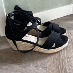 Svarta kilklack sandaler med snörning - Snygga svarta sandaler med kilklack och korsade remmar. Skorna har snörning runt vristen och en plattformsula i repimitation. Perfekta för sommaren och ger en stilren look till alla outfits. Inköpta på Din Sko för 1 år sedan. Gått i dem en gång sen har de bara stått i garderoben. Nypris 500:-. Eventuell lite stor i storlek, har vanligtvis 37/38 men dessa passar mig. Längd från häl till tå: 23-24cm
