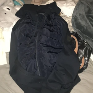 Mörkblå cardigan från Polo Ralph Lauren - Mörkblå cardigan från Polo Ralph Lauren med dragkedja framtill och broderad logotyp på ärmen. Jackan har quiltad framsida och huva, vilket ger en sportig och stilren look. Perfekt för lager-på-lager under kyligare dagar.