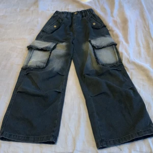 Svarta cargopants med stora fickor - Säljer ett par svarta cargopants med bred passform och stora fickor på sidorna. Byxorna har fransiga kanter på ficklocken, resår i midjan och knappdetaljer. Perfekta för en avslappnad och cool stil. Fickorna längst upp där fram är inte riktiga. Det är Y2K fashion 😎