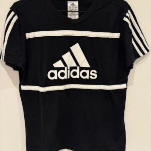 Svart t-shirt från Adidas med vita detaljer - Svart t-shirt från Adidas med klassiska vita ränder på axlarna och två vita band över bröstet. Stor Adidas-logga framtill. Perfekt för en sportig look och enkel att matcha med jeans eller träningsbyxor.