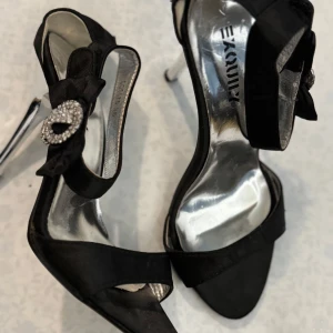 Svarta klackskor med rosett och strassdetalj - Säljer ett par eleganta svarta klackskor från Exquily med öppen tå, stor rosett och glittrande strassdetalj på sidan. Skorna har smal klack och en stilren design som passar perfekt till festliga tillfällen.