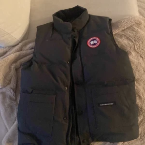 Canada gosse och så har den bra kvalitet  - Svart dunväst från Canada Goose med klassisk logotyp på bröstet och stora fickor framtill. Västen har hög krage och knäppning med både dragkedja och knappar. Perfekt för lager-på-lager under kyliga dagar.