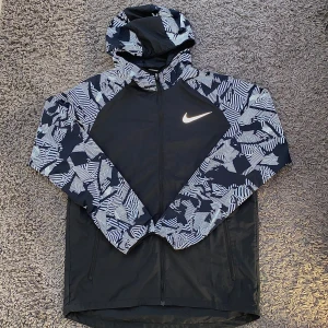 Svart vindjacka - Nike vindjacka med svart bas och vita grafiska mönster på ärmar och huva. Jackan har hel dragkedja, två fickor och huva. Perfekt för löpning eller utomhusaktiviteter. Snygg och sportig design med Nike-logga på bröstet.