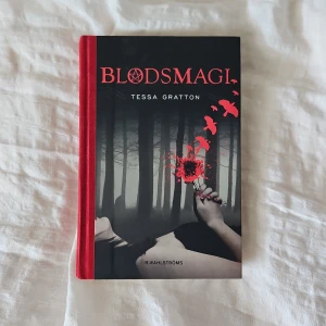 Blodsmagi - Blodsmagi av Tessa Gratton är en mörk fantasybok med hård pärm och röd rygg. Omslaget visar en hand som håller en blodröd blomma i en dimmig skog, med röda fåglar som flyger. Boken handlar om magi, blod och hemligheter.