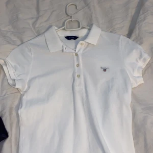 Vit piké t-shirt från GANT - Klassisk vit piké t-shirt från GANT med krage och knappar framtill. T-shirten har korta ärmar och broderad GANT-logga på bröstet. Perfekt för en stilren och sportig look.