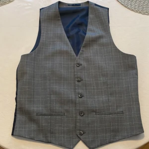 Grå rutig väst från River Island - Snygg grå väst från River Island med diskret rutigt mönster och fem knappar framtill. Västen har två fickor fram och justerbart spänne i ryggen. Perfekt för att lyfta din outfit med en klassisk touch.