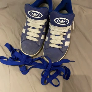 Blå Adidas Campus sneakers med vita ränder - Säljer ett par blå Adidas Campus sneakers med klassiska vita ränder och gummisula. Skorna har rund tå, platt sula och kommer med både vita och blå skosnören. Perfekta för dig som gillar retrostil och streetwear. 9/10 det enda som skulle behövas göras är att tvätta upp lite.