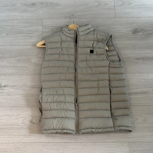 Ljusgrå dunväst från The Boston Vest - Säljer en ljusgrå dunväst från The Boston Vest med hög krage och dragkedja framtill. Västen har en stilren, quiltad design och är perfekt för lager-på-lager. Passar dig som vill ha en enkel och snygg look. Perfekt nu till sommaren