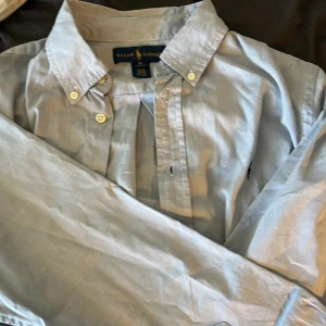 Ljusblå skjorta från Ralph Lauren - Klassisk ljusblå skjorta från Ralph Lauren med button-down krage och broderad logga på bröstet. Skjortan har långa ärmar och knäppning framtill. Perfekt för en stilren och tidlös look.