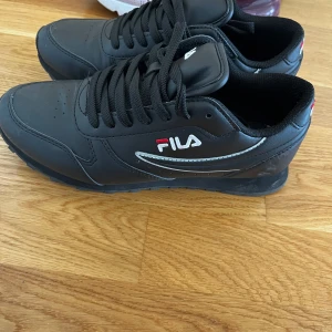 Svarta sneakers från Fila - Säljer ett par svarta Fila sneakers med klassisk design och svarta skosnören. Skorna har rund tå, platt sula och Fila-logga på sidan och baksidan. Perfekta för en sportig och stilren look. Storlek 37