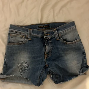 Blå jeansshorts från Nudie Jeans - Säljer ett par klassiska blå jeansshorts från Nudie Jeans med fransig kant och slitning på vänster framsida. Shortsen har fem fickor, knappgylf och snygga detaljer på bakfickorna.