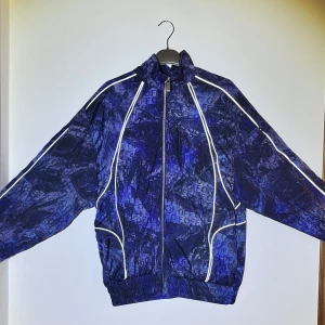 Blå mönstrad vindjacka från Off-White - Säljer en blå och svart vindjacka från Off-White med unikt mönster och vita detaljer längs dragkedjan och ärmarna. Jackan har hög krage, hel dragkedja och elastisk midja. Perfekt för dig som vill sticka ut med streetwear.