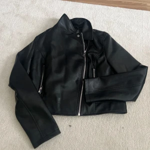 Svart skinnjacka med dragkedja - Säljer en svart skinnjacka i croppad modell med klassisk krage och dragkedja framtill. Jackan har långa ärmar och två fickor med dragkedja. Perfekt för en cool och stilren look.