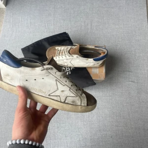 Vita och bruna sneakers från Golden Goose - Golden Goose sneakers i vitt skinn med bruna mockadetaljer på tån och blå häl. Klassisk stjärndesign på sidan och snörning framtill. Snygg vintage-look och passar perfekt till streetwear.5/10 i skick. Du får med dig allt som finns på bilden 