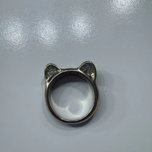 Ring med kattöron och stenar - Säljer en unik ring i silverfärg med söta kattöron och små glittriga stenar på öronen. Perfekt för dig som gillar djur eller vill ha något annorlunda i din smyckessamling. Ringen har en slät och blank yta.