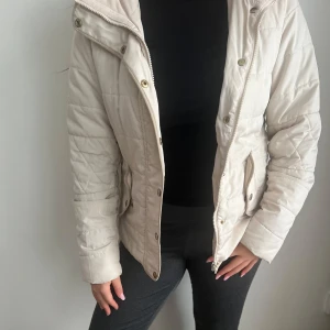 Beige quiltad jacka med dragkedja och knappar - Säljer en beige quiltad jacka med både dragkedja och knappar framtill. Jackan har hög krage och långa ärmar, perfekt för kyligare dagar. Snygg och enkel att matcha till olika outfits.