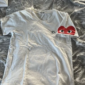 Vit t-shirt från Comme des Garçons - Säljer en vit t-shirt från Comme des Garçons med det ikoniska röda hjärtat med ögon på ärmen. T-shirten har en rund halsringning och korta ärmar. Perfekt för en stilren och trendig look.