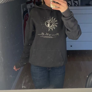 Mörkgrå hoodie med sol- och måntryck - Säljer en svart hoodie med unikt tryck av sol och måne framtill och ett stort månfas-mönster på ryggen. Tröjan har huva och magficka, banden till luvan är borttappade.