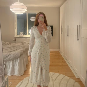 Vit långklänning från Adoore - Superfin vit långklänning från Adoore med små blå blommor, v-ringning och knappar framtill. Klänningen har långa ärmar och markerad midja för en feminin siluett. Perfekt för sommarens tillfällen! Använd enstaka gång så är precis som ny💗 