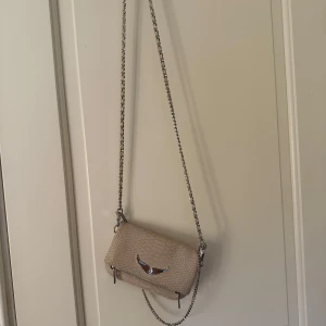 Zadig väska  - Snygg beige axelväska med ormmönstrad struktur och silverfärgad kedjerem. Väskan har ett dekorativt metallspänne framtill och stängs med dragkedja. Perfekt storlek för det viktigaste och ger en trendig touch till din outfit.