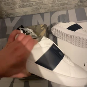 Vita sneakers med mörkblå detalj - Säljer ett par stilrena vita sneakers med en bred mörkblå remsa på sidan. Skorna har rund tå och platt sula, perfekt för en clean och modern look.