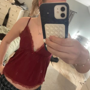 Vinröd linnetopp med spets från Nelly - Säljer en vinröd linnetopp från Nelly med tunna axelband, spetsdetaljer längs urringningen och nederkanten samt en liten rosett framtill. Toppen är mjuk och har en feminin look, perfekt för sommaren.