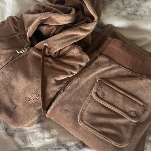 Brun velour hoodie och byxor  från Juicy Couture - Mjuk och lyxig brun hoodie och byxor   från Juicy Couture med dragkedja och broderad JC-logga framtill. Perfekt för en avslappnad och trendig look. Klassisk huva och fickor framtill. Som nya!