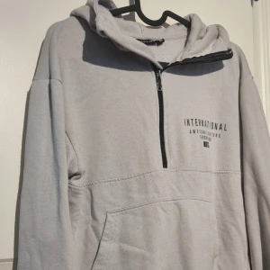 Ljusgrå hoodie från Reaction med tryck - Säljer en ljusgrå hoodie från Reaction med svart tryck på bröstet och ärmen. Tröjan har huva, halv dragkedja och en stor magficka. Perfekt för en avslappnad stil och enkel att matcha med jeans eller joggers.