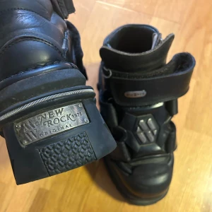 Svarta boots från New Rock - Säljer ett par svarta boots från New Rock med kraftig plattformssula och metallplatta på hälen. Skorna har spännen och detaljer i metall samt en robust design som sticker ut. Perfekta för dig som gillar alternativ stil.