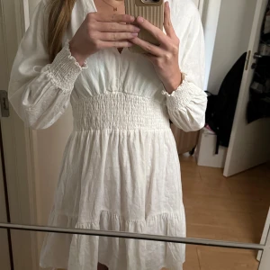 Vit klänning - Säljer en söt vit klänning med v-ringad hals och långa ärmar med smock vid handlederna ifrån Zara! Klänningen har smock i midjan och volang nertill, vilket ger en fin siluett. Perfekt för sommarens alla tillfällen eller till student!🥂Klänningen är i storlek S