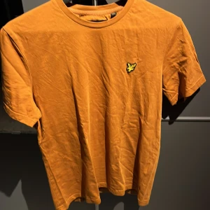 Gul t-shirt från Lyle & Scott str S - Säljer en gul t-shirt från Lyle & Scott med klassisk rund hals och korta ärmar. På bröstet finns en broderad gul logotyp i form av en fågel. T-shirten är i mjuk bomull och har en enkel, stilren design. (NY)