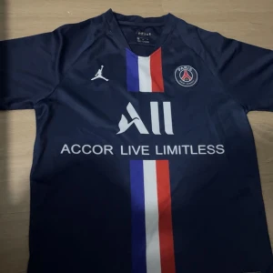 PSG fotbollströja från Jordan - Mörkblå PSG fotbollströja från Jordan med klubbmärke på bröstet och vit Jordan-logga. Vertikal rand i blått, vitt och rött framtill. Tillverkad i lätt och ventilerande material, perfekt för fotbollsträning eller match.