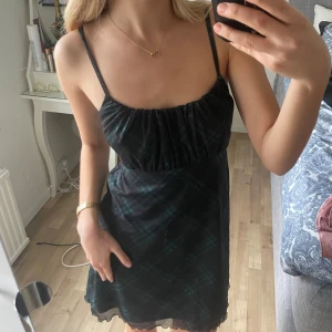 Svart och grön rutig kortklänning med smala axelband - Supersöt svart kortklänning med grönt rutmönster och smala axelband. Klänningen har en lätt rynkad byst och sitter bekvämt med normal passform. Perfekt för sommardagar eller när du vill känna dig extra fin.