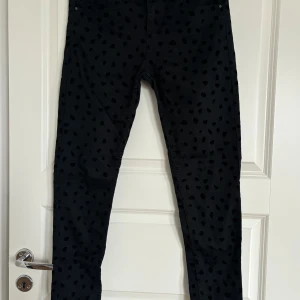 Svarta byxor med mönster från H&M - Svarta byxor från H&M med svarta prickar som mönster. Klassisk femficksmodell med knapp och dragkedja framtill. Byxorna har en slim passform och är tillverkade i ett stretchigt material för extra komfort.