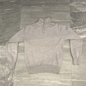 Ljusgrå stickad tröja från Stenströms - Säljer en stilren ljusgrå stickad tröja från Stenströms med hög krage och tre knappar i olika färger vid halsen. Tröjan har lång ärm och ribbade muddar vid ärmslut och nederkant. Perfekt för en clean och klassisk look.