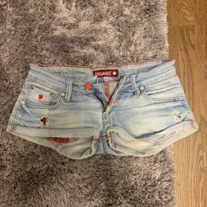 Ljusa jeansshorts från Dsquared2 - Säljer ett par ljusblå jeansshorts från Dsquared2 med röda detaljer och slitningar framtill. Shortsen har klassisk femficksdesign, röd broderad lönnlöv på fickan och röd knapp. Perfekta för sommaren! Skriv privat om mått 