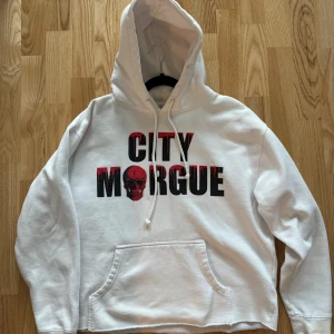 Vit hoodie City Morgue x VLONE - Vit hoodie med tryck 'CITY MORGUE' och röd dödskalle framtill. Stor röd V och illustration av två svarta hundar på ryggen. Klassisk känguruficka och huva med dragsko. Perfekt statement-plagg för streetwear.