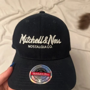 Svart keps från Mitchell & Ness - Snygg svart keps från Mitchell & Ness med broderad logga framtill och justerbar passform. Klassisk böjd skärm och detaljer i vitt och rött på insidan. Perfekt accessoar för en sportig look.