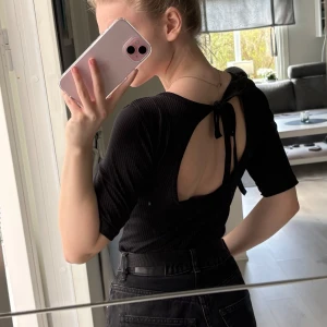 Svart ribbad topp med öppen rygg - Svart ribbad topp med korta ärmar och snygg öppen rygg med knytband i nacken. Perfekt för dig som gillar stilrena och trendiga plagg med en twist.☺️
