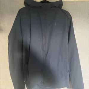 Blå vindjacka under armour - Säljer en blå vindjacka från Under Armour med huva och dragkedja framtill. Strl M sällsynt modell hör av er vid frågor