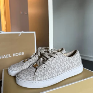 Beige sneakers från Michael Kors  - Snygga beige sneakers från Michael Kors med MK-logga över hela skon. Skorna har vita sulor, snörning med gulddetaljer och är tillverkade i syntetmaterial. Perfekta för dig som gillar stilrena och trendiga skor.