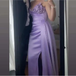 Lila långklänning med paljettdetaljer - Säljer en elegant ljuslila långklänning med glittriga paljetter upptill och en snygg slits framtill. Klänningen har omlottliv och axelbandslös design, perfekt för bal eller fest. Materialet är glansigt och ger en lyxig känsla.