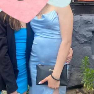 Ljusblå långklänning från NELLY - Säljer en elegant ljusblå långklänning från NLY med smala axelband och draperad urringning. Klänningen har en slits framtill och är tillverkad i ett glansigt satinliknande material. Perfekt för bal eller festliga tillfällen.