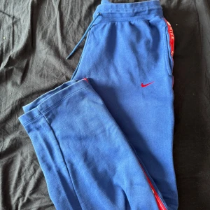 Blå mjukisbyxor från Nike med röda detaljer - Säljer ett par blå mjukisbyxor från Nike med röda detaljer och tryckta loggor längs sidorna. Byxorna har snörning i midjan och tryckknappar längs benen. Perfekta för en avslappnad stil. Finns en fläck på byxorna jag ej får bort. 