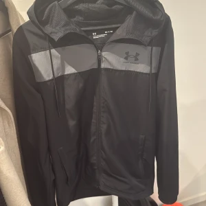 Svart vindjacka från Under Armour - Säljer en svart vindjacka från Under Armour med grå panel över bröstet och logga på vänster sida. Jackan har huva, dragkedja framtill och två sidofickor. Perfekt för träning eller vardag.