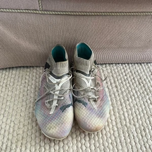 Vita puma future - Säljer ett par snygga fotbollsskor från Puma i vitt med inslag av pastellfärger och svarta detaljer. Skorna har rosa dobbar och snörning framtill. Perfekta för dig som vill sticka ut på planen!