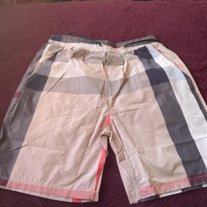 Burberry swim shorts - Snygga beige shorts med stora rutor i svart, vitt och rött. De har elastisk midja med snörning och en bakficka. Perfekta för sommaren och har en avslappnad passform.
